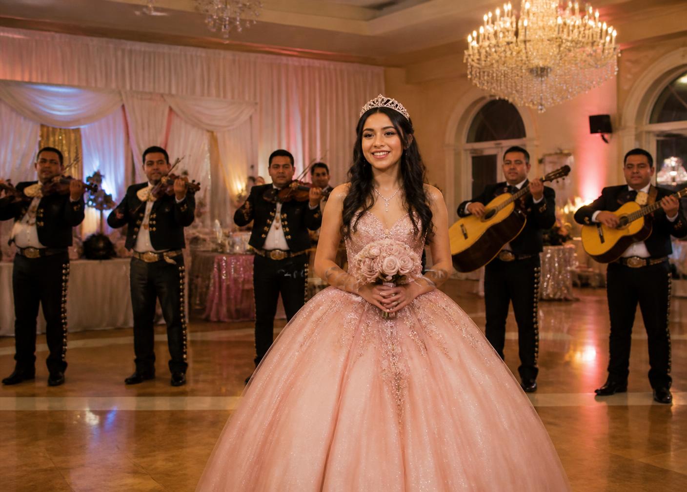 Quinceañera sonriendo en su fiesta con músicos de mariachi al fondo.